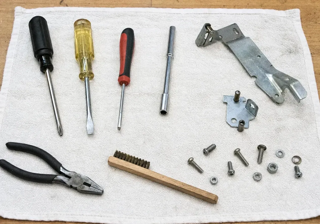 Four screwdrivers, pliers and a wooden-handled wire brush arranged on a cloth with a hinge bracket, bolts and nuts